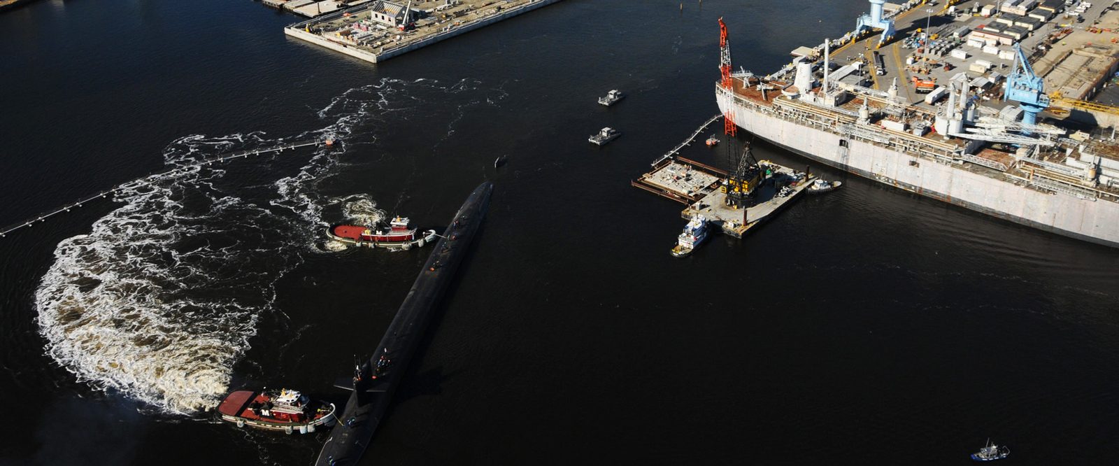 Aerial view of the Norfolk Naval Shipyard in Portsmouth, Virginia, with a submarine under way flanked by tugboats